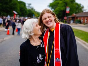 A young woman in an academic gown poses with an elderly loved one outside a university graduation ceremony.