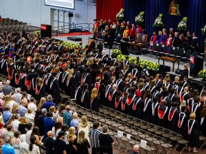 A large group of people in academic robes stand in front of their chairs at a university graduation while an audience looks on.