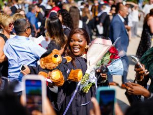A woman in a Convocation gown who is holding a teddy bear and flowers celebrates outside while people take photos of her with their phones.