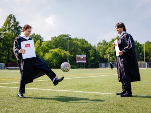 Two young men in academic robes kick a soccer ball while holding their diplomas outside a university graduation ceremony.