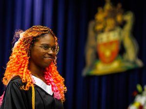 A young woman in an academic robe smiles on stage at a university graduation ceremony.