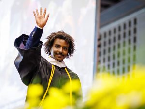 A young man in an academic robe smiles and waves from the stage at a university graduation ceremony.