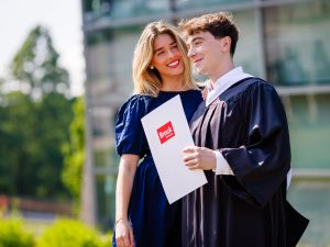 A young man in an academic robe holds a diploma and poses for a photo with a loved one.