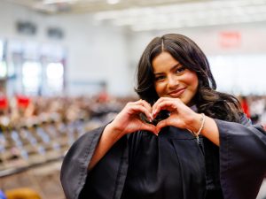 A woman in a Convocation gown makes a heart sign with her hands.