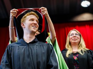 A young man in an academic robe stands while someone places a ceremonial sash over his shoulders at a university graduation ceremony.