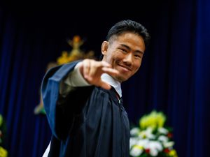 A young man in a academic robe smiles and raises his hand while on stage at a university graduation ceremony.