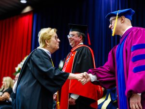 Two people in academic robes shake hands on stage at a university graduation ceremony.