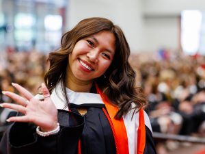 A young woman with brown hair wearing academic robes smiles and waves at the camera during a convocation ceremony.
