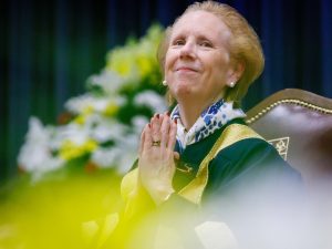 A woman in academic robes sits on an elaborate chair with yellow and white flowers in the background and smiles with her hands in a thankful gesture.