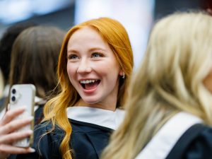 A young woman with red hair and wearing academic robes stands in line waiting for the convocation ceremony to begin. She’s smiling and holding up her phone.