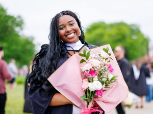 A smiling young woman with long braided black hair wears academic robes and holds pink and white flowers in front of trees under a blue sky.