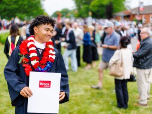 A young man with brown curly hair stands outdoors with a crowd behind him. He’s wearing academic robes, two red and white floral lei, and holds a Brock University degree folder.