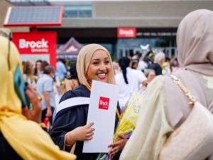 A young woman wearing academic robes and a beige Hijab smiles as she holds a Brock University degree folder. She is outdoors with a crowd and a Brock sign in the background.