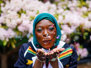 A young woman wearing academic robes and a turquoise traditional headscarf blows confetti from her hands towards the camera in front of a floral background.