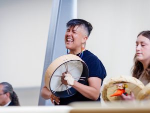 A trio of Indigenous drummers performs during a convocation ceremony. A seated man is on the left, and the two women, in centre and on the right, are drumming.