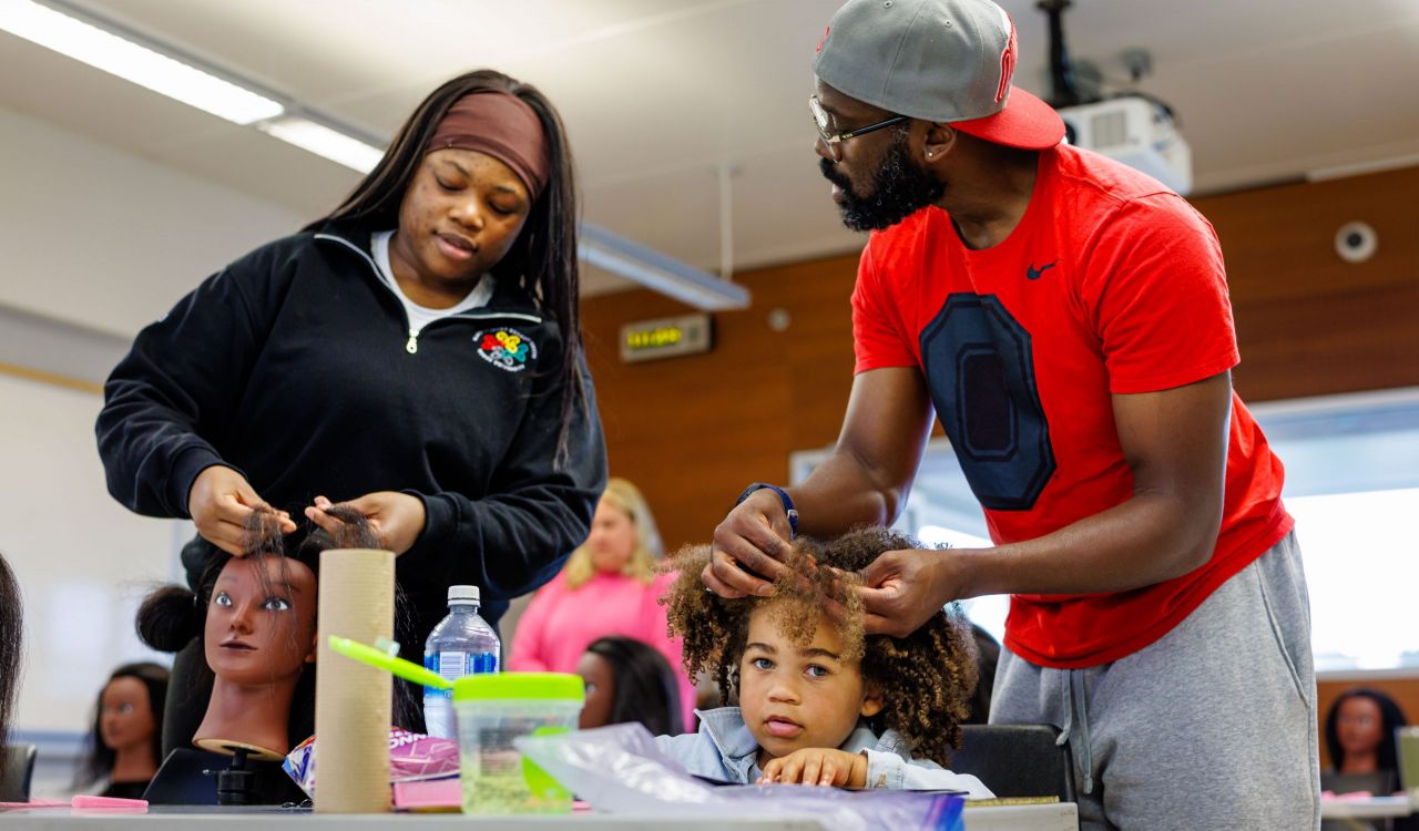 A man carefully braids a young child’s hair while seated at a table during a hands-on wellness activity. Hair tools and supplies are visible in the foreground as the child looks upward.
