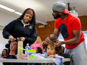 A man carefully braids a young child’s hair while seated at a table during a hands-on wellness activity. Hair tools and supplies are visible in the foreground as the child looks upward.