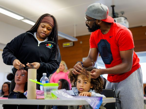A man carefully braids a young child’s hair while seated at a table during a hands-on wellness activity. Hair tools and supplies are visible in the foreground as the child looks upward.