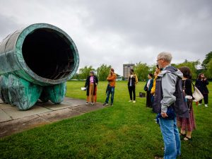 Outdoors under a slightly grey sky, a group stands around looking at a large sculpture while a tour guide provides information about the piece of art.