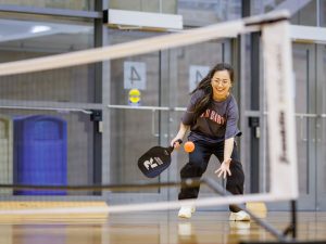 A woman smiles in a gymnasium while holding a paddle and attempting to catch a ball.