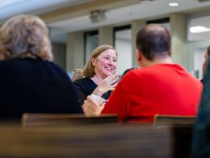 In a cafeteria setting, a woman smiles and speaks animatedly to the group sitting around her. One person in the group is holding a spoon and a white container of ice cream