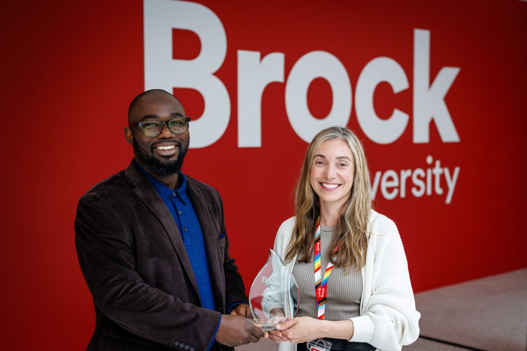 A man and a woman stand in front of a red wall with the Brock University logo, smiling for the camera, and holding a glass trophy between them.