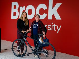 Two women stand in front of a red wall, with a Brock University logo on it. Both women are smiling, looking at the camera, and holding on to a bicycle.