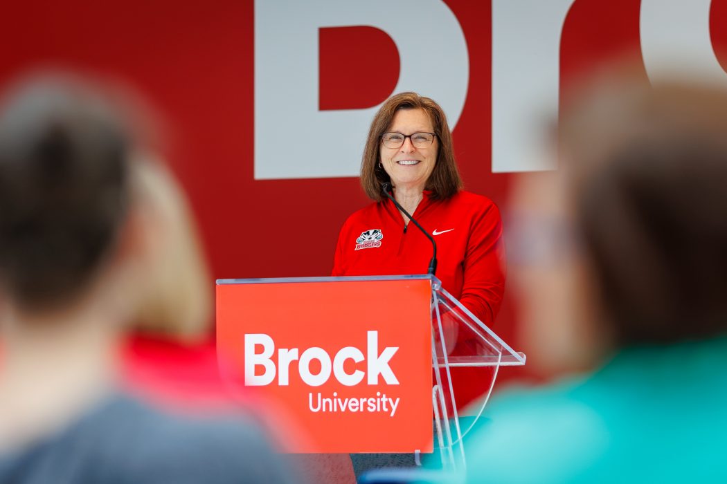 A woman stands at a podium that features a Brock University logo, in front of a red wall, and smiles at the crowd she is addressing.