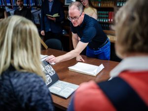 In a library setting, a man speaks to a group of people as he flips the page in a photo album. The book, sitting on a table, features black and white archival photos.