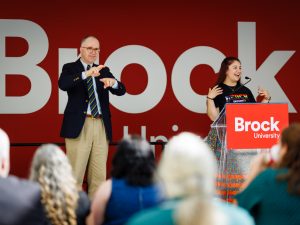 A young woman dressed in a floral indigenous ribbon skirt stands at a podium and animatedly addresses the crowd. To her left is a gentleman providing American Sign Language interpreting. Behind them both is a red wall featuring the Brock logo.