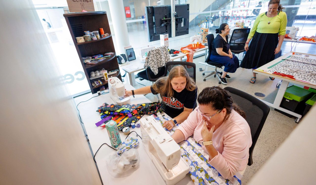 Four women are gathered in a craft room setting surrounded by fabrics, ribbon and sewing equipment. Two women in the foreground are seated at a sewing machine, sewing white ribbon on floral fabric. Two women are behind animatedly talking.