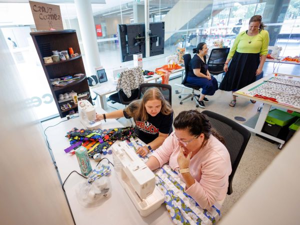 Four women are gathered in a craft room setting surrounded by fabrics, ribbon and sewing equipment. Two women in the foreground are seated at a sewing machine, sewing white ribbon on floral fabric. Two women are behind animatedly talking.