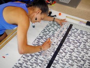 A woman leans over a crafting table covered with fabric featuring a forest print. With one of her hands, she’s holding a ribbon in place on the fabric, and with her other hand she’s using a glue stick to adhere the ribbon to the fabric.