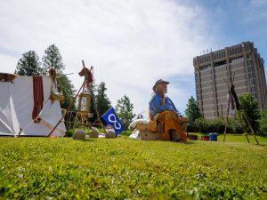 A man sits on a bench wearing Indigenous beaded garments and accessories. Behind him is a Métis encampment display featuring numerous items including a tent, flag, pelts and clay pots. The Brock University tower is seen in the background.