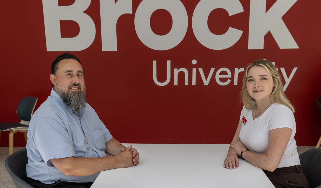 Two people sit facing each other at a small round table in a bright indoor campus space. Behind them, a large red wall displays a white Brock University logo. Both individuals are seated on modern chairs with their hands resting on the table and are smiling at the camera.