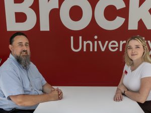 Two people sit facing each other at a small round table in a bright indoor campus space. Behind them, a large red wall displays a white Brock University logo. Both individuals are seated on modern chairs with their hands resting on the table and are smiling at the camera.