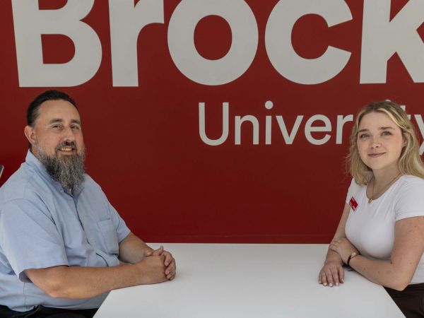Two people sit facing each other at a small round table in a bright indoor campus space. Behind them, a large red wall displays a white Brock University logo. Both individuals are seated on modern chairs with their hands resting on the table and are smiling at the camera.