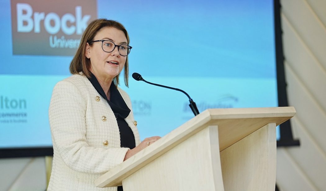 A woman speaks into a microphone at a podium with a presentation screen behind her.