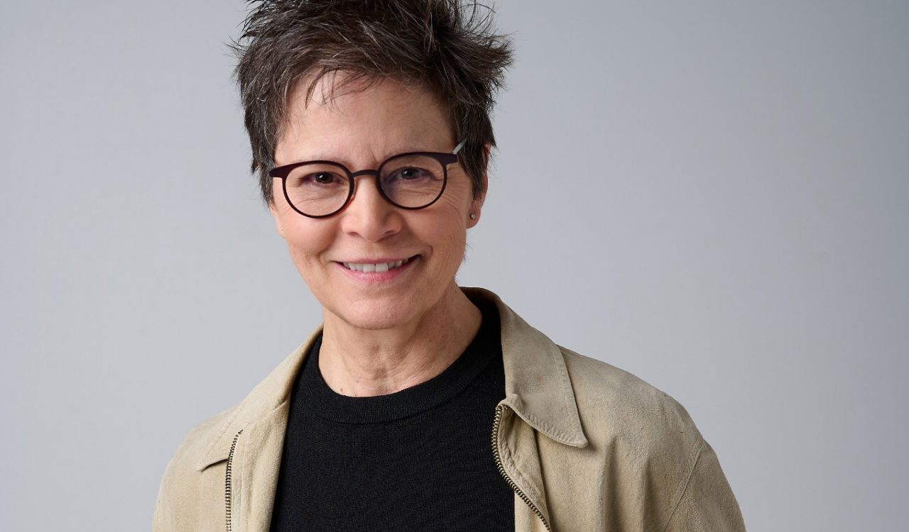 A woman stands smiling with her hands in her pockets.