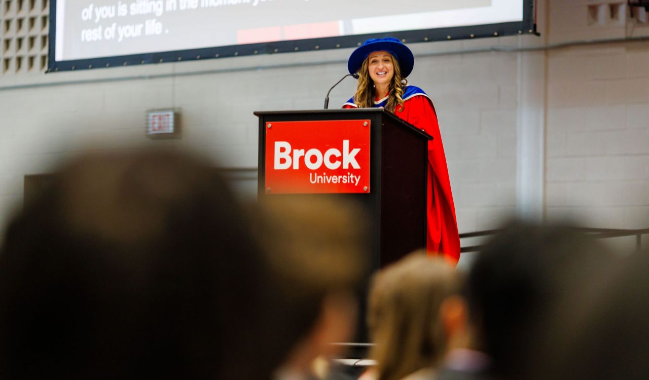 A woman in academic regalia speaks from a podium during a university graduation ceremony.