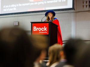 A woman in academic regalia speaks from a podium during a university graduation ceremony.