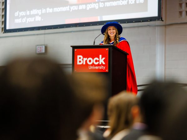 A woman in academic regalia speaks from a podium during a university graduation ceremony.