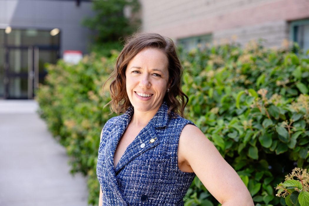 Woman in blue dress stands in front of a green bush and brown building displaying two pins on her lapel. One a Beta Gamma Sigma pin the other a Brock alumni pin.