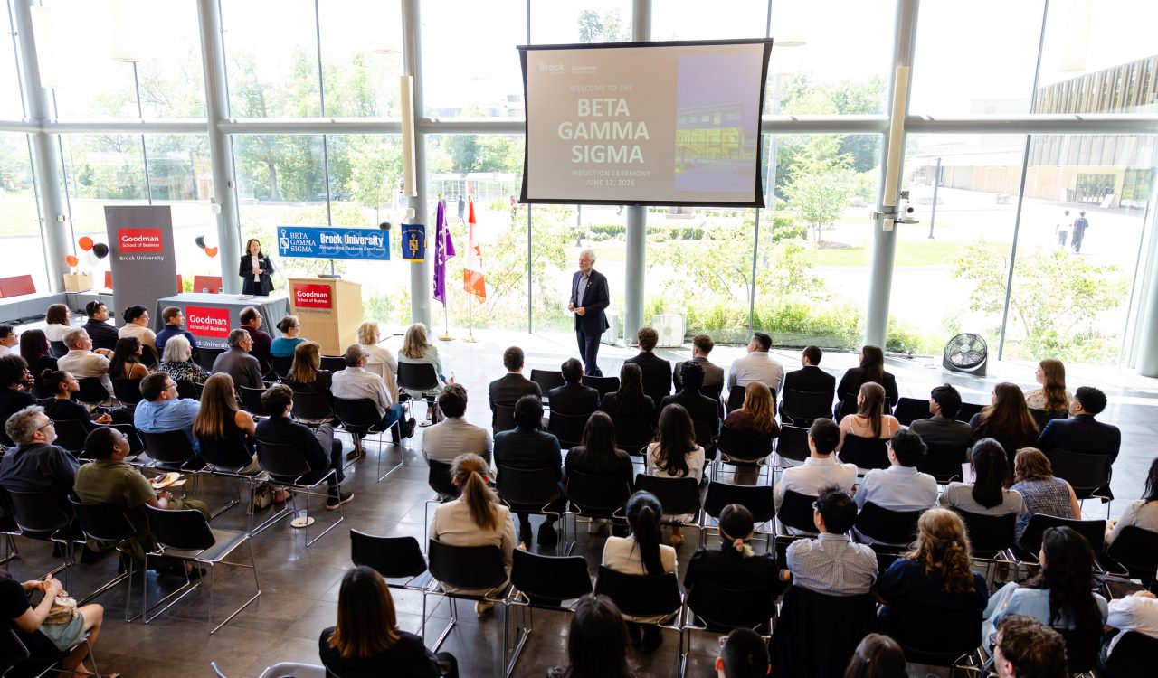 In a large glass framed room a man stands in front of 120 full chairs speaking to the audience.