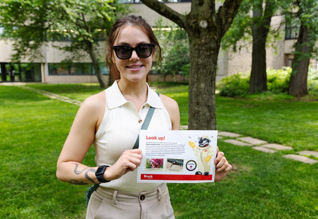 A woman stands outside holding a sign that talks about Brock's designation as a Bee Campus.