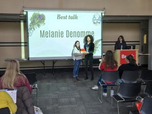 Two people stand together with an award between them at the front of a room.
