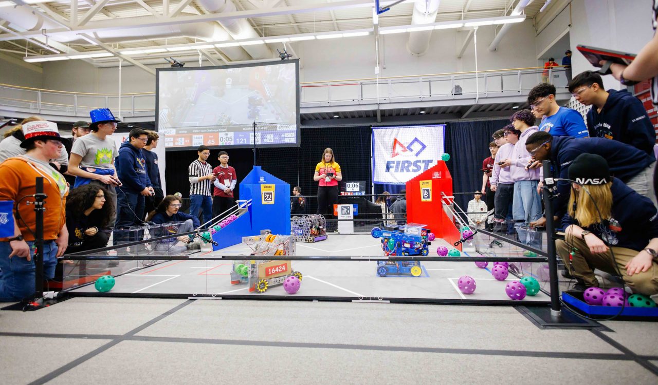 A group of people control robots that are seen moving around in a marked ring.