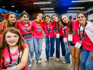 A group of girls in pink team t-shirts pose in a semi-circle at a robotics competition.
