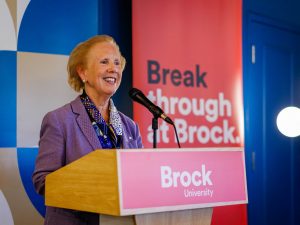 A woman stands at a podium speaking into a microphone. A Brock University-branded banner stands in behind her.