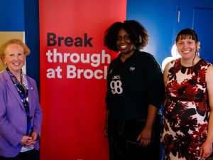 Three woman stand together surrounding a Brock University banner that reads Break through at Brock.